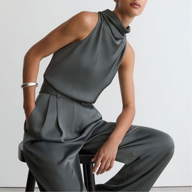 Model wearing Reiss dark grey sleeveless halterneck top and wide-leg trousers, seated on a stool, styled with a silver cuff bracelet.