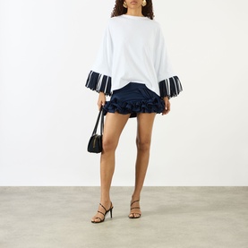 Model wearing Jacquemus navy mini skirt with ruffled hem, styled with white fringed-sleeve top and black heeled sandals, carrying black bag