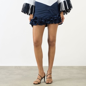 Model wearing Jacquemus navy mini skirt with ruffle hem detail, styled with a fringed top and strappy heeled sandals.