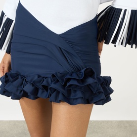 Model wearing Jacquemus navy mini skirt with crossover waist and tiered ruffle hem, styled with a white long-sleeve top with fringe cuffs.