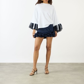 Model wearing Jacquemus navy mini skirt with ruffled hem, styled with a white fringed-cuff top and strappy heeled sandals.
