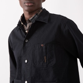 Model wearing Nudie Jeans Black Casual Jacket in cotton with chest pocket, styled over a brown plaid shirt.
