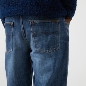 Model wearing Nudie Jeans Rad Rufus Straight-Leg Jeans in Blue Twisted Tracks, showing rear pockets with tobacco stitching and logo patch detail.