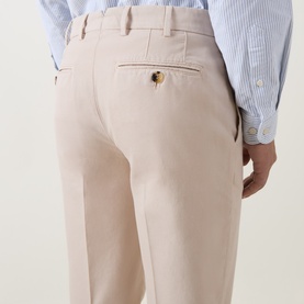 Model wearing Brunello Cucinelli sand cotton chino trousers, rear view showing back pocket with gold button detail, styled with a blue striped shirt.