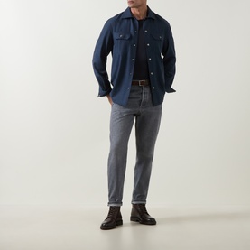 Model wearing Brunello Cucinelli grey cotton jeans, styled with a navy overshirt and dark brown leather boots.
