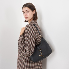 Model carrying Strathberry Kite Sling Crossbody Bag in black grained leather with gold-tone hardware, worn over shoulder, styled with houndstooth