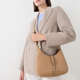 Model carrying Strathberry Kite Sling Crossbody Bag in caramel grained calf leather, gold-tone Music Bar hardware, worn over shoulder. Styled with