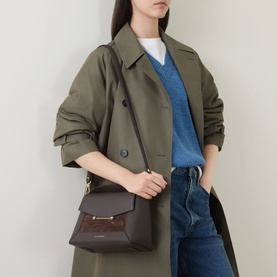 Model carrying Strathberry Mosaic Top Handle Bag in Dark Chocolate grained leather, gold-tone hardware, worn crossbody with olive trench coat.