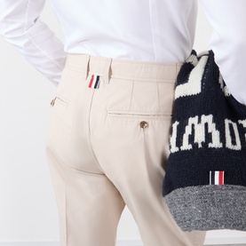 Model wearing Thom Browne Tapered Pressed-Crease Chinos in beige, showing rear jet pockets with signature RWB tab detail, styled with navy knitwear.