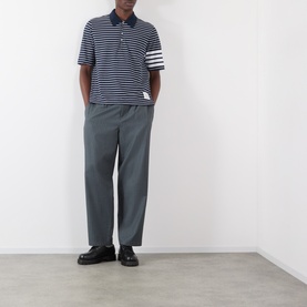 Model wearing Thom Browne navy and white striped cotton polo with 4-bar sleeve detail, styled with grey tailored trousers and black loafers.