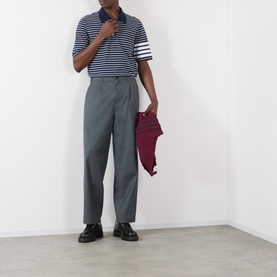 Model wearing Thom Browne navy/white stripe cotton polo with grey tailored trousers and black loafers, carrying burgundy jacket.