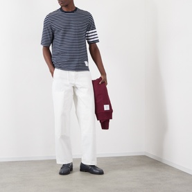 Model wearing Thom Browne navy and white striped cotton T-shirt with four-bar detail, styled with white trousers and carrying a burgundy jacket.