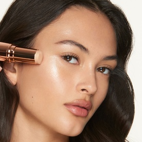Model with Charlotte Tilbury Unreal Highlighter Stick in Glazed Goddess applied to cheekbone, rose gold packaging visible