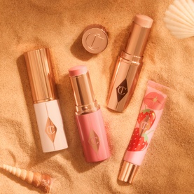 Charlotte Tilbury Unreal Highlighter Stick in Glazed Goddess, shown with matching blush sticks and lip gloss on sandy beach backdrop
