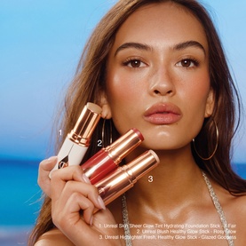 Model with Charlotte Tilbury Unreal Highlighter Stick in Glazed Goddess, shown with Healthy Glow Stick and Foundation Stick against beach backdrop