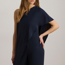 Model wearing Lauren navy one-shoulder jumpsuit with draped cape sleeve, styled with a gold bangle bracelet.