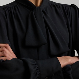 Model wearing Lauren black shirt with oversized bow neckline, ruched sleeves, and button-front detail, arms crossed.