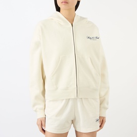 Model wearing Sporty and Rich ecru cotton zip-up hoodie with script logo, styled with matching shorts.
