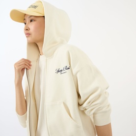 Model wearing Sporty and Rich ecru zip-up cotton hoodie with embroidered logo, styled with a cream cap.