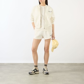 Model wearing Sporty & Rich Ecru cotton hoodie and matching shorts, styled with grey socks, chunky trainers, holding a cap.