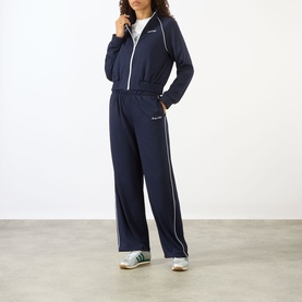 Model wearing Sporty and Rich dark navy zip-up track jacket with white piping, styled with matching wide-leg trousers and green trainers.
