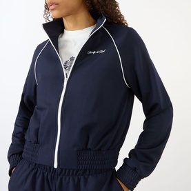 Model wearing Sporty and Rich dark navy zip-up track jacket with white piping and logo, styled with matching navy trousers and white top.