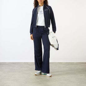 Model wearing Sporty & Rich dark navy track jacket with white piping, styled with matching wide-leg trousers, white tee and green trainers.