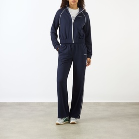 Model wearing Sporty and Rich Dark Navy/White wide-leg track pants with white piping, styled with matching zip-up track jacket and green trainers.