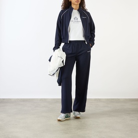 Model wearing Sporty and Rich Dark Navy/White track trousers and matching zip-up jacket, styled with a white graphic tee and grey trainers.