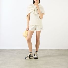 Model wearing Sporty and Rich ecru cotton shorts with matching top and bow detail, styled with grey trainers and carrying a small yellow bag.
