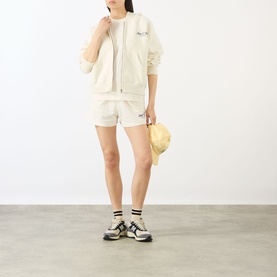 Model wearing Sporty and Rich ecru cotton shorts, styled with matching zip jacket, white tee, trainer socks and chunky trainers, holding a sun hat.