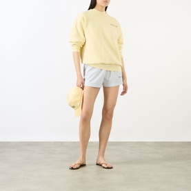 Model wearing Sporty & Rich Almond Navy cotton sweatshirt with white shorts and tan cap, styled with brown flip-flops