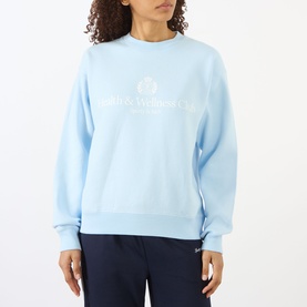Model wearing Sporty and Rich Ciel blue crew neck sweatshirt with white logo print, styled with navy trousers