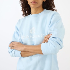 Model wearing Sporty and Rich Ciel sweatshirt with embroidered logo graphic, arms crossed, cropped close-up view