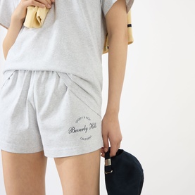 Model wearing Sporty and Rich grey cotton shorts with Beverly Hills logo print, styled with a grey t-shirt. Model holds a navy cap.