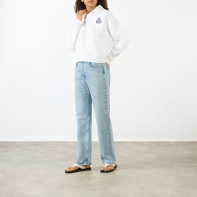 Model wearing Sporty and Rich white sweatshirt with small chest graphic, styled with light blue straight-leg jeans and black flip-flops.