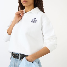 Model wearing Sporty and Rich white cropped polo-collar sweatshirt with navy crest embroidery, styled with light blue jeans and black belt.