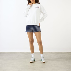 Model wearing Sporty and Rich white sweatshirt with small chest graphic, styled with navy striped shorts and white trainers.