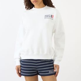Model wearing Sporty and Rich white crew neck sweatshirt with chest logo graphic, styled with navy striped shorts