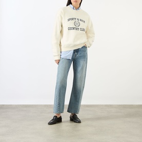 Model wearing Sporty and Rich ecru cotton sweatshirt with varsity logo, styled with light blue jeans and black loafers.