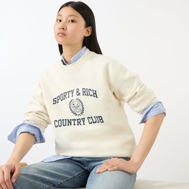 Model wearing Sporty and Rich ecru cotton crew-neck sweatshirt with navy Country Club logo graphic, styled with a blue striped shirt and jeans.