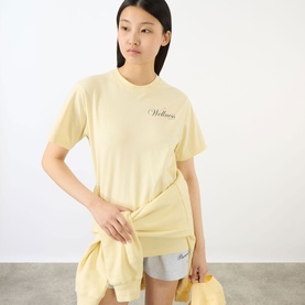 Model wearing Sporty and Rich Almond Navy cotton T-shirt with script logo, styled with matching shorts and yellow jacket tied at waist.