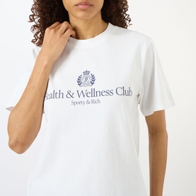 Model wearing Sporty and Rich white oversized T-shirt with navy Health & Wellness Club crest graphic print to chest.