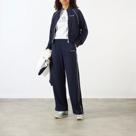 Model wearing Sporty and Rich white T-shirt styled with navy track jacket, wide-leg trousers with white piping, and green trainers.