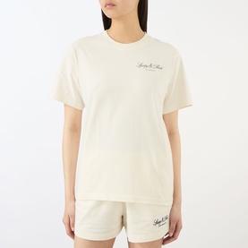 Model wearing Sporty and Rich ecru cotton T-shirt with script logo, styled with matching ecru shorts.