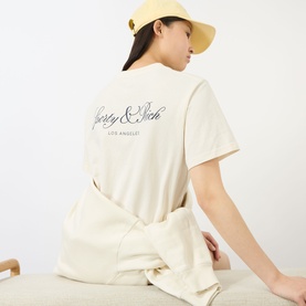Model wearing Sporty and Rich ecru cotton T-shirt, back view showing script logo print, styled with matching ecru trousers and yellow hat