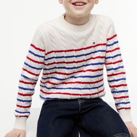 Model wearing Tommy Hilfiger cable-knit cream jumper with red and blue stripes, paired with dark navy jeans.