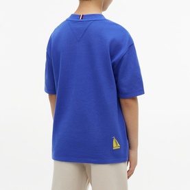 Model wearing Tommy Hilfiger Noble Blue short-sleeve sweatshirt, rear view, with small sailboat logo patch, styled with beige trousers.