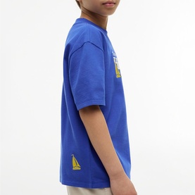 Model wearing Tommy Hilfiger Noble Blue kids T-shirt, side view showing yellow sailboat graphic on hem and chest print.