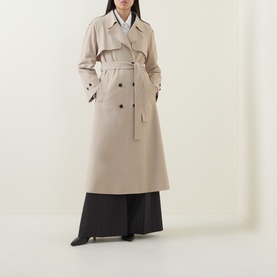 Model wearing HIGH Smuggler Trench Coat in beige; double-breasted, belted longline style with notch lapel, styled with black wide-leg trousers
